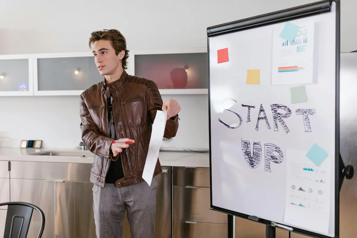 Young startup founder standing in a modern office kitchen, holding a financial report and presenting a "Start Up" strategy on a whiteboard with data charts.
