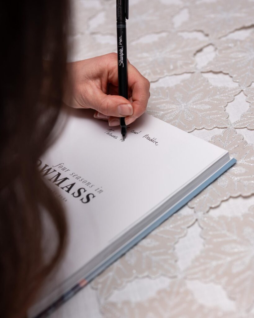 Close-up of a hand writing a personal inscription inside a book on a lace-covered table