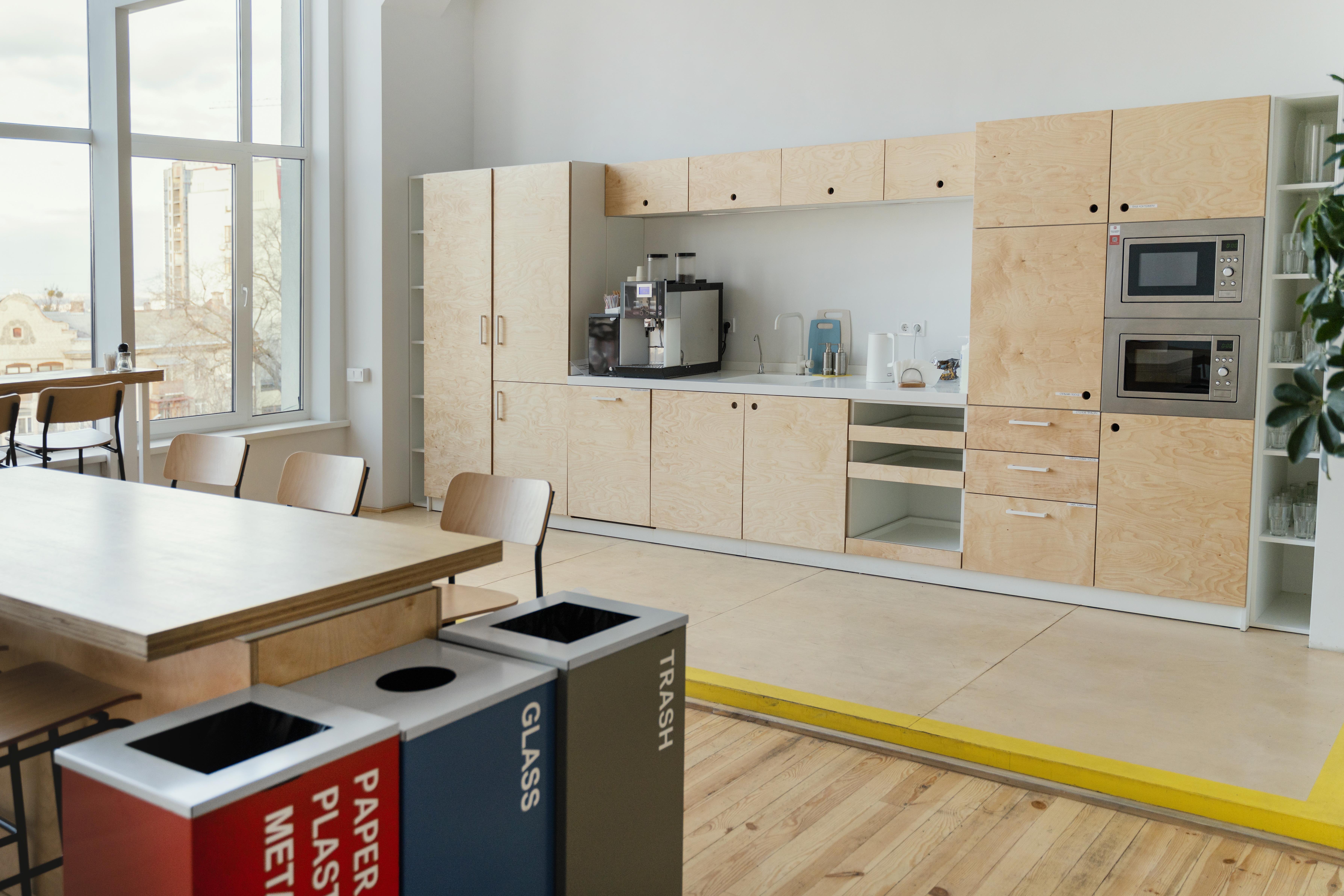 Modern office kitchen with light wood modular hybrid shelf cabinets, coffee machine, microwave, large windows, recycling bins, and seating. Bright, organised space.