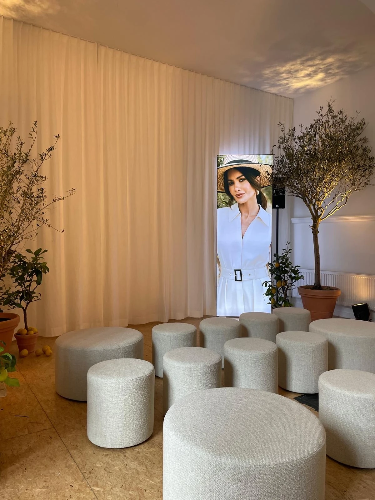 Presentation corner with beige poufs, olive trees and portrait screen between cream curtains.