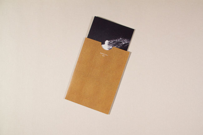 A light brown cardboard folder lies flat on a light beige surface. The folder is open, revealing a black photo inside. The photo depicts a white, vaguely humanoid shape against a dark background with visible white streaks or markings. The folder’s flap is folded over, partially obscuring the photo.
