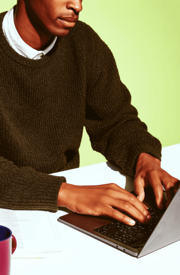 A person typing on a laptop, wearing a green sweater, on a white table with a red cup in the background.