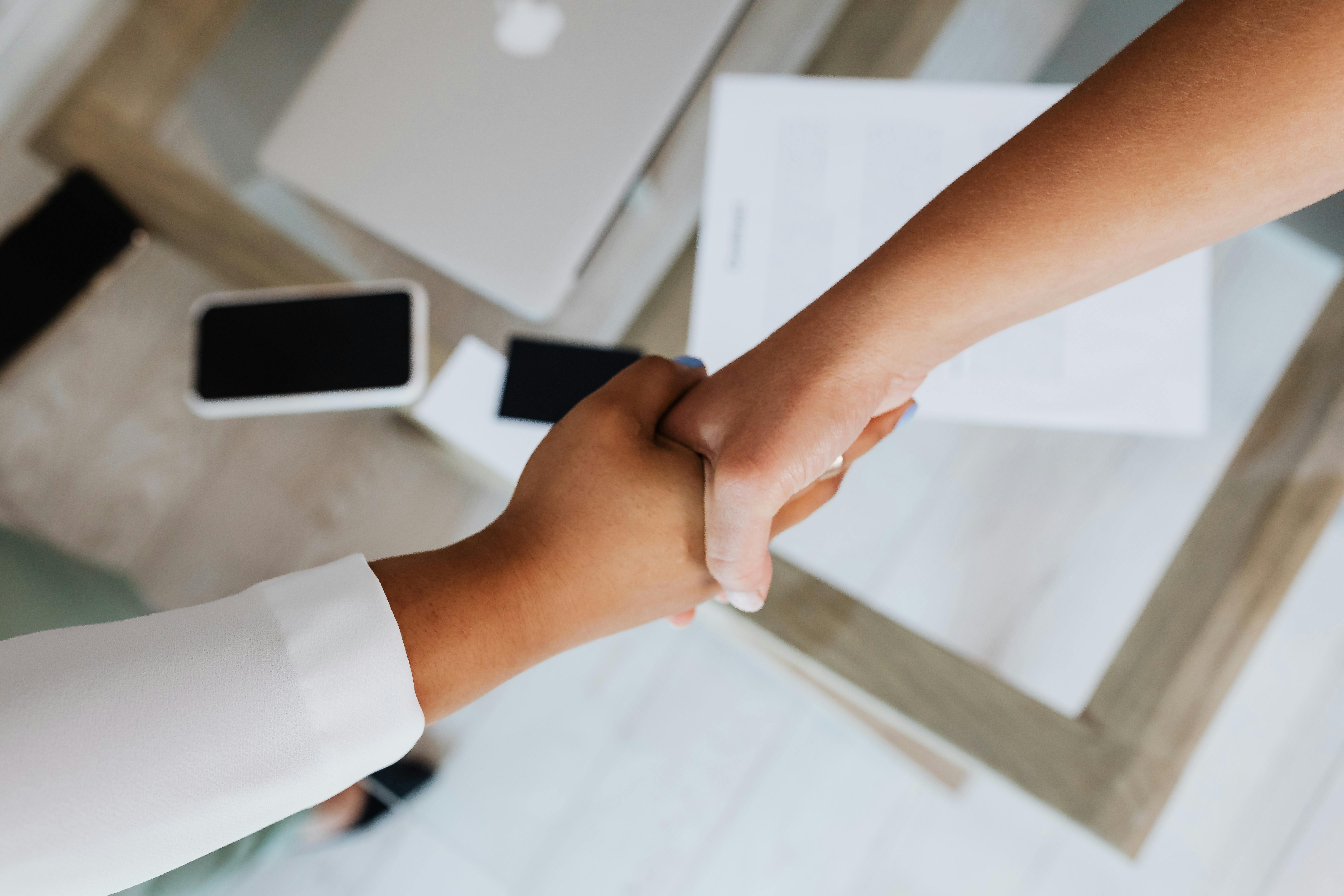 Two hands shaking over a desk with a laptop and documents, symbolizing a agreement of mutual promotion