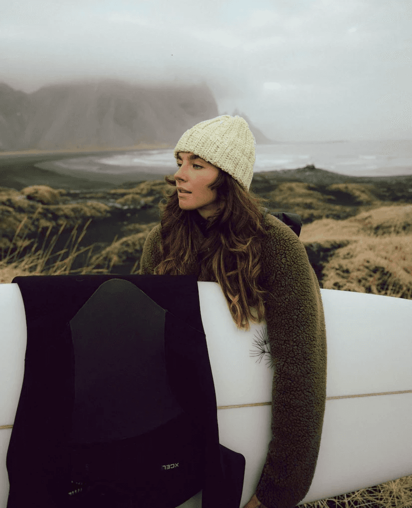 A person in a cozy brown sweater and knit hat holds a surfboard standing in front of a foggy, rugged coastal landscape with distant mountains and grey skies, capturing an adventurous spirit in a serene natural setting.