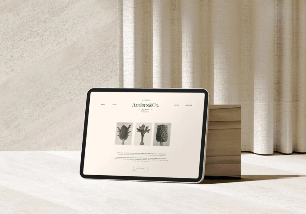 Horizontal tablet mockup displaying a minimalist website design with neutral tones, propped against a wooden block. Set on a light textured surface with soft curtain background.