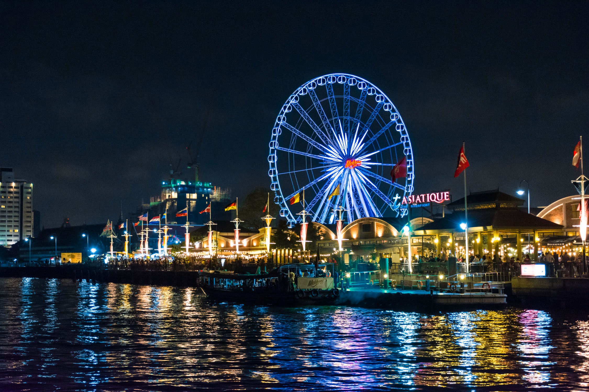 A lit Ferris wheel and carnival rides at a waterfront pier at night with colorful lights reflecting on the water.