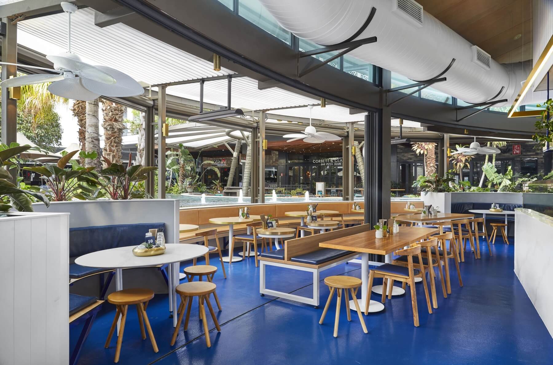 Modern restaurant interior design with bright blue flooring, custom wooden booth seating, exposed ceiling ductwork, and views of an outdoor shopping precinct.