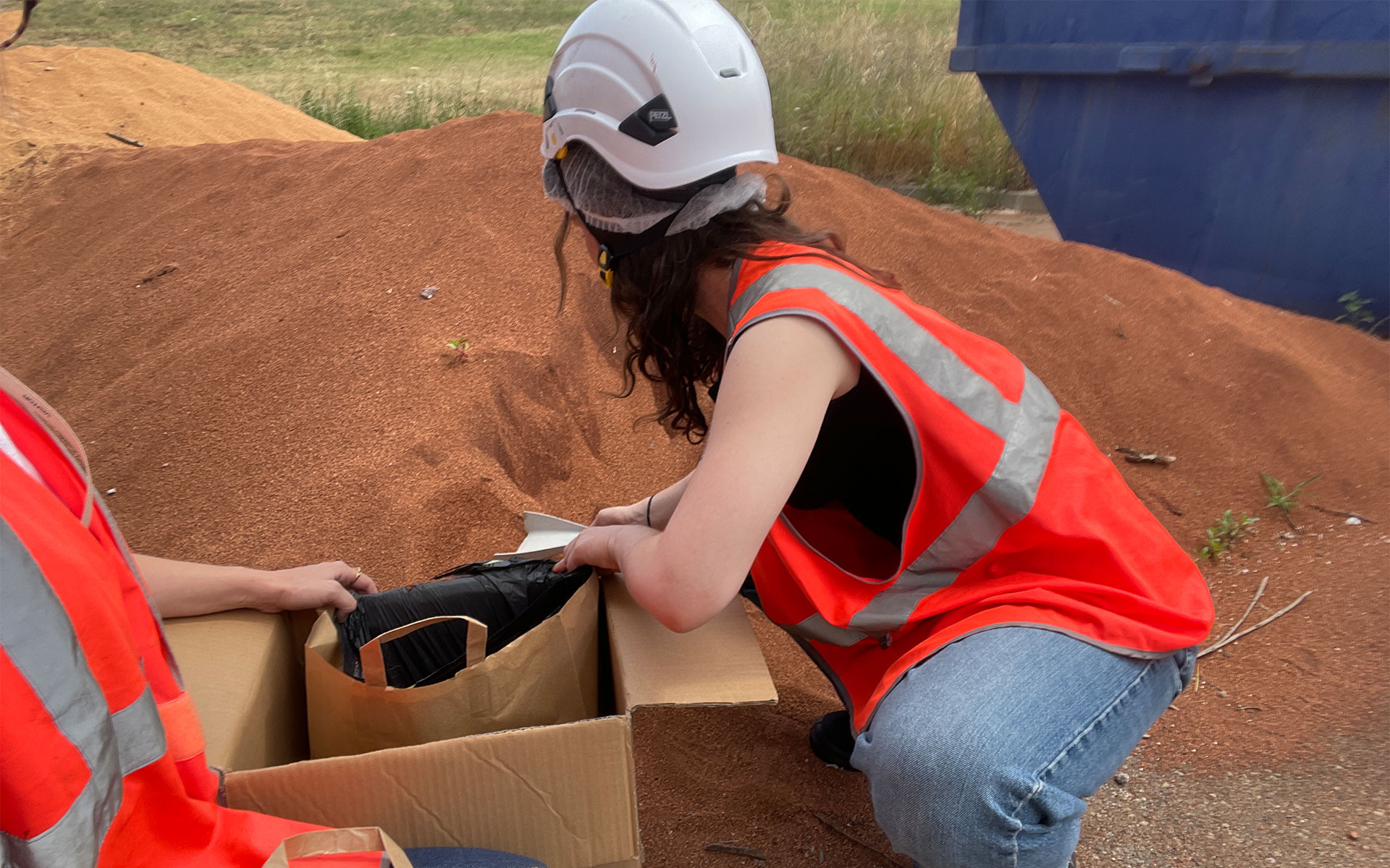 Deux personnes portant des gilets de sécurité orange réfléchissants travaillent à l'extérieur, accroupies près d'un grand tas de terre rouge. La personne au premier plan, portant un casque de chantier blanc, range un sac en papier brun contenant un sac poubelle noir dans une boîte en carton. Une benne bleue est visible en arrière-plan, suggérant un environnement de chantier ou de gestion des matériaux.