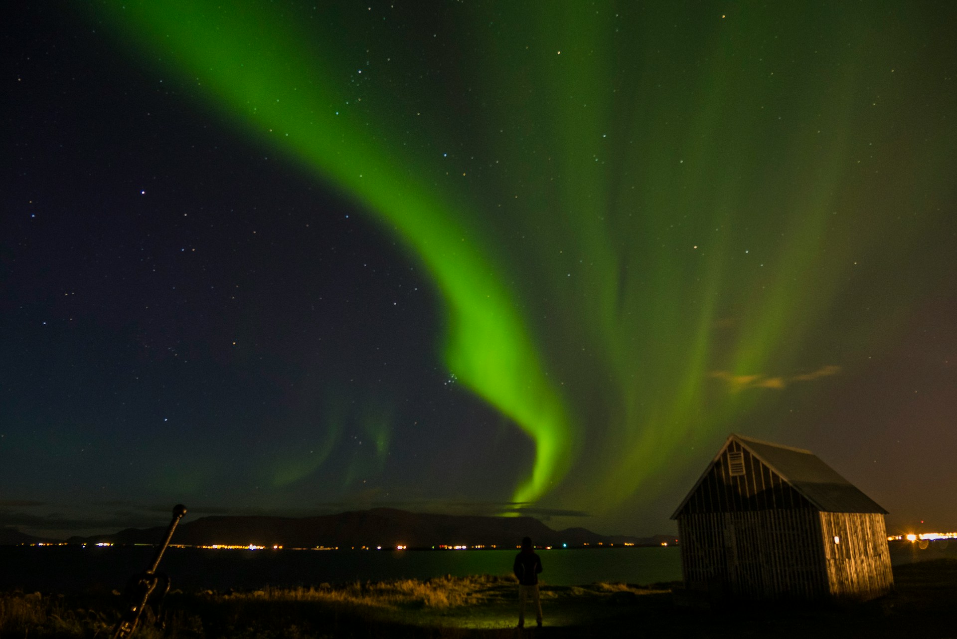 Green northern lights illuminate the night sky above Reykjavík's coastline with a small cabin in the foreground.