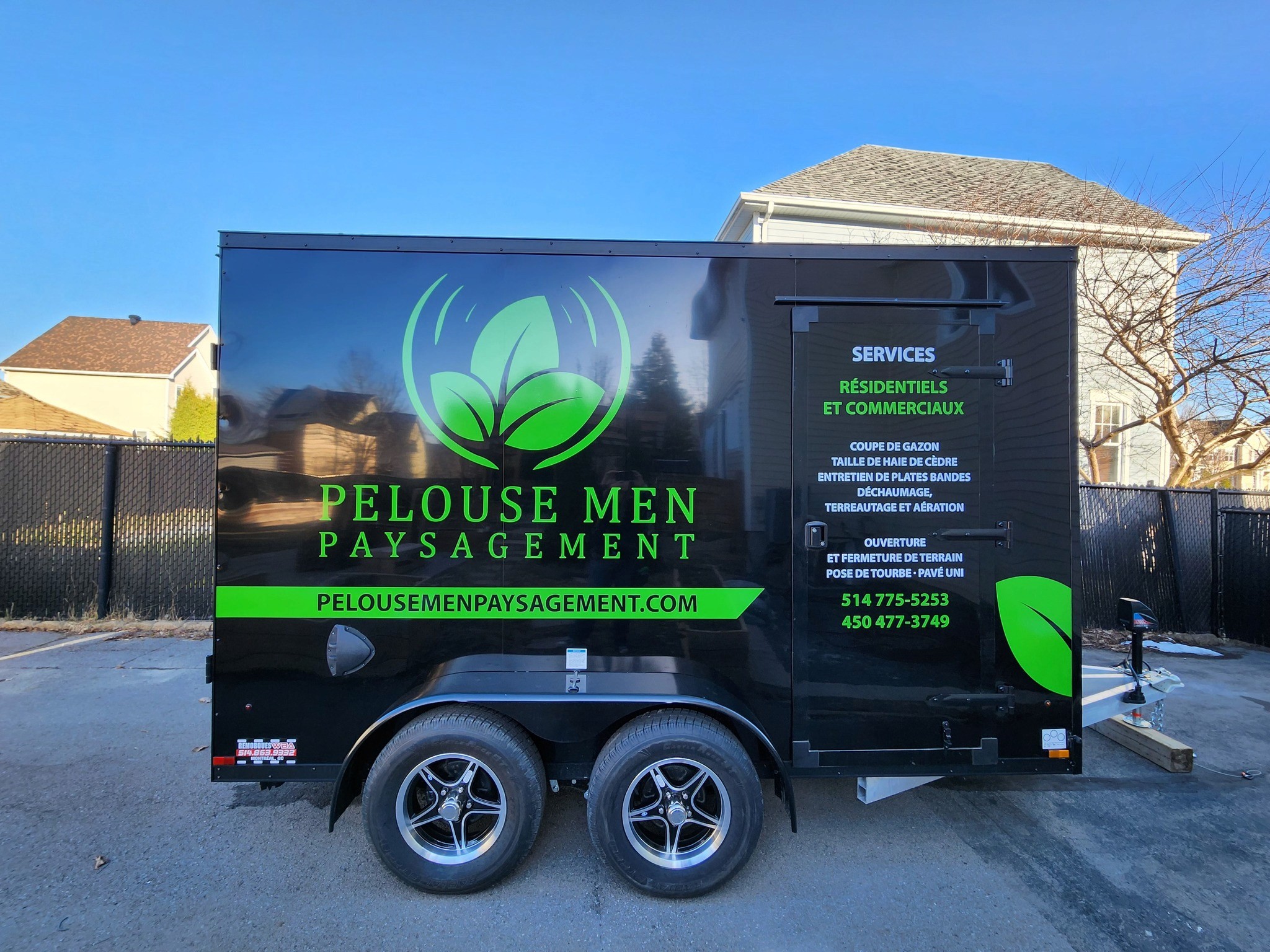 Remorque noire Pelouse Men Paysagement avec lettrage vert et logo de feuille, stationnée dans une entrée résidentielle.