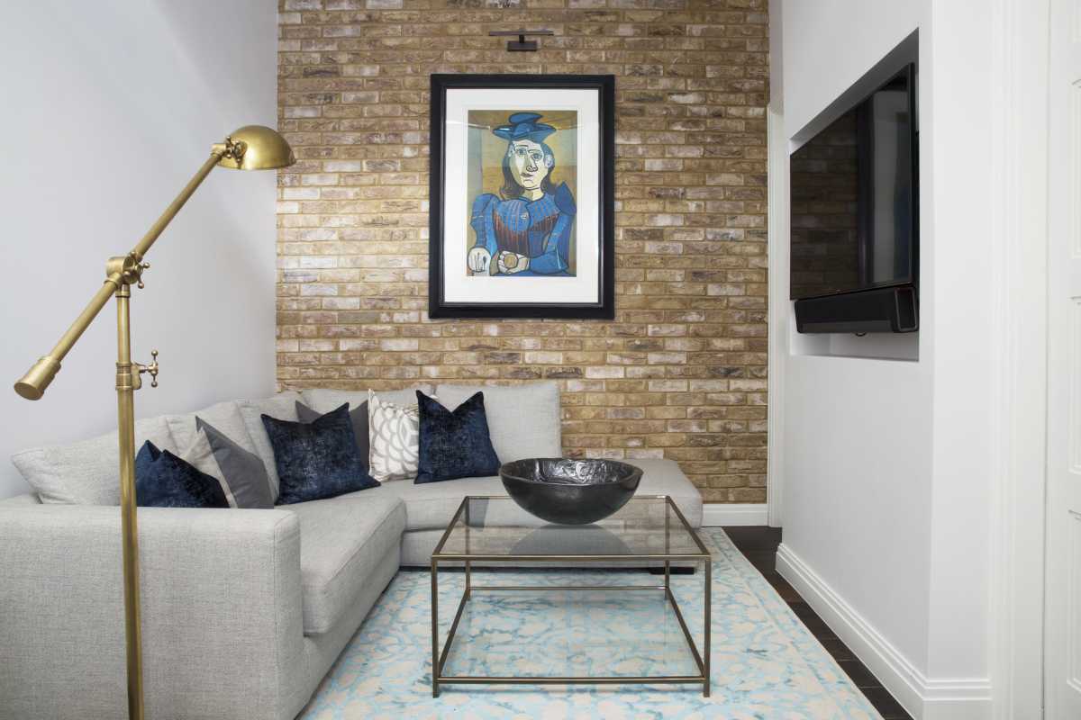 Cozy, modern living room styled by interior designers in Lymington, featuring an exposed brick feature wall, light grey sectional sofa, and framed art piece.