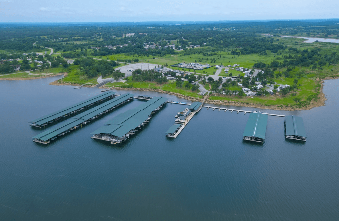 Aerial view of a large keystone harbor marina on a calm, expansive keystone lake, featuring several long docks with green-roofed boat slips, surrounded by lush greenery and distant land.