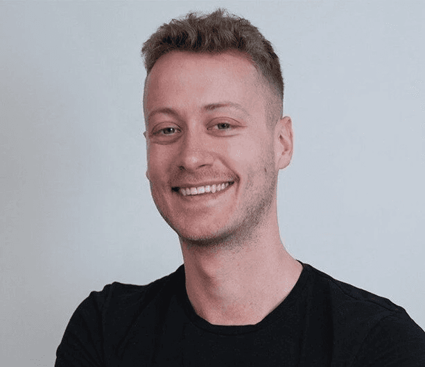 Professional headshot of a smiling young man with short blonde hair wearing a black t-shirt against a plain white background.