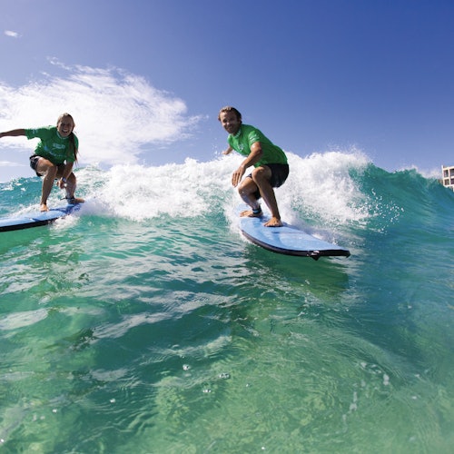 Zwei Personen surfen auf blauen Surfboards, tragen grüne Shirts und reiten eine Welle unter einem klaren blauen Himmel.