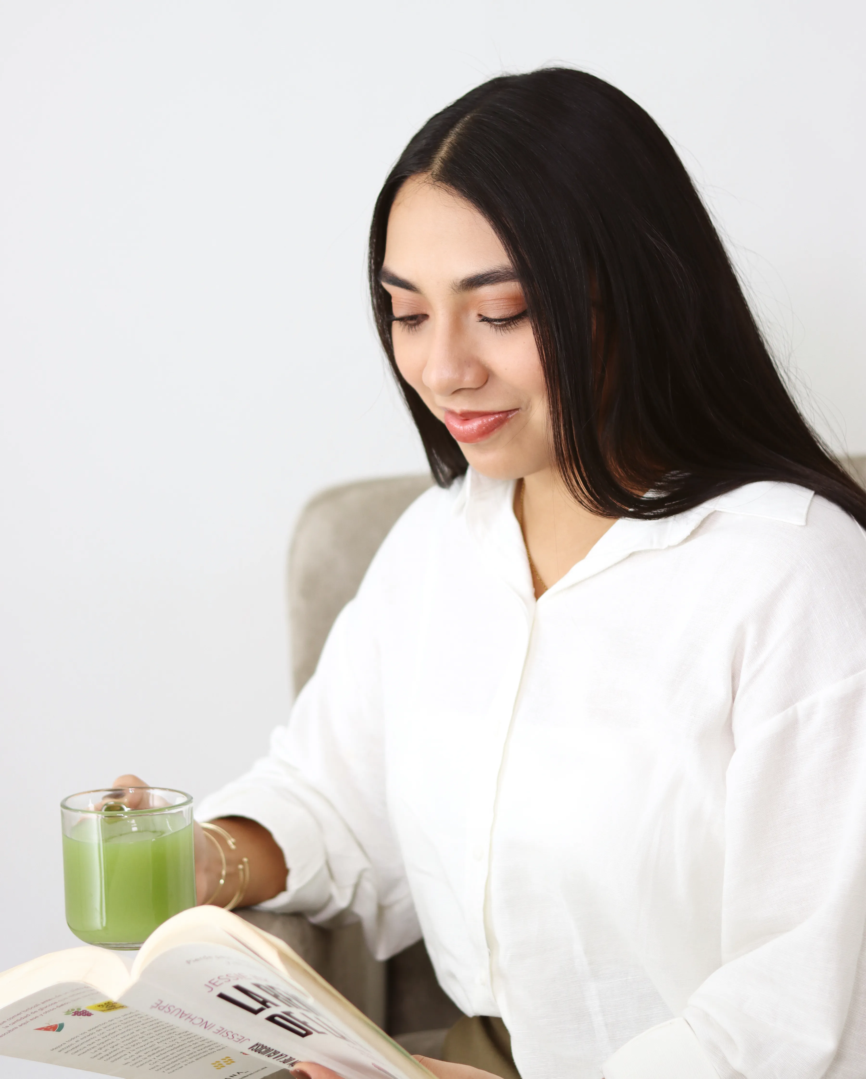 Mujer con camisa blanca sosteniendo bebida verde mientras lee.