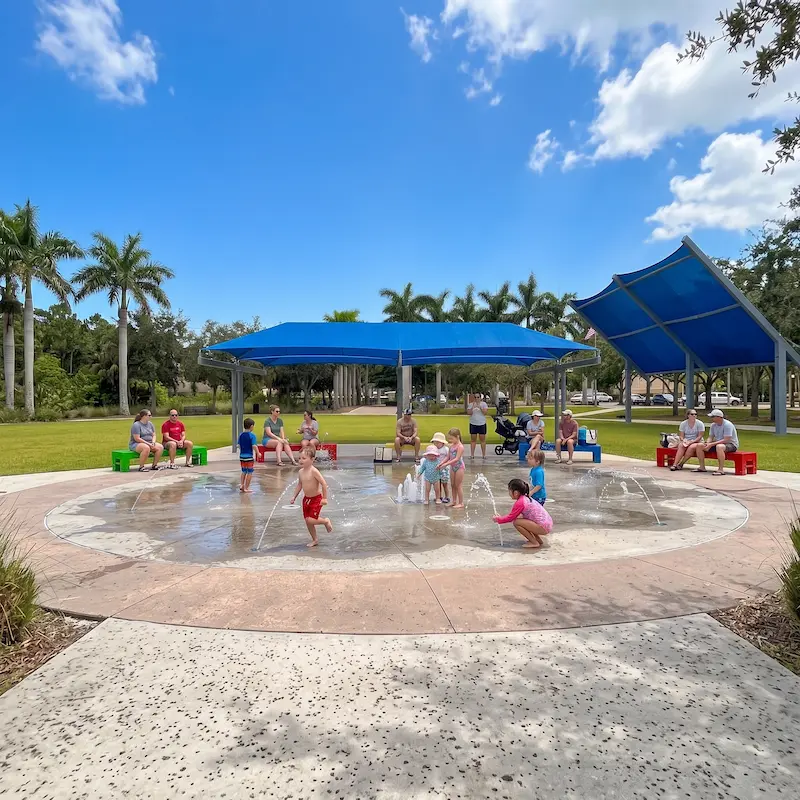 A vibrant splash pad with colorful water jets and blue shade structures under a partly cloudy sky is surrounded by lush greenery and palm trees along the Riverwalk in Bradenton, FL.