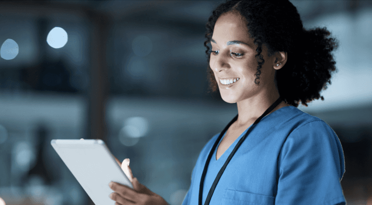 A nurse wearing glasses and blue scrubs smiling while looking at a tablet or clipboard in a healthcare setting