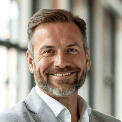 a professional headshot of a smiling man wearing a light gray suit jacket and a white shirt.