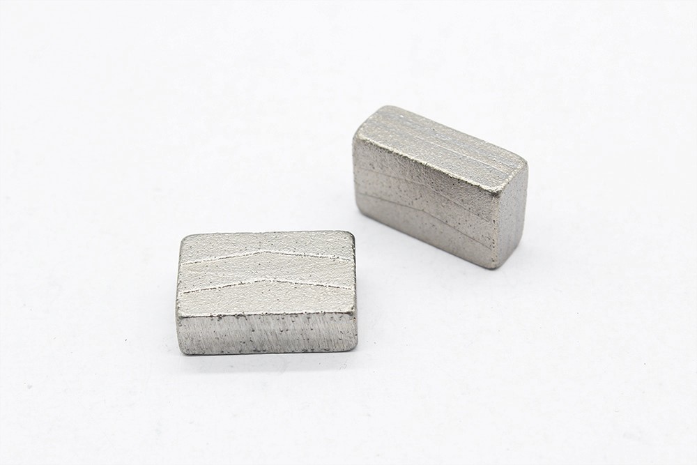 Two rectangular diamond segment with silver white gloss on the surface and V-shaped patterns on the sides are placed on a white background tabletop. They are used for granite cutting