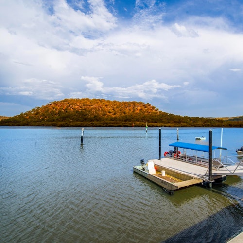 一个湖岸上停泊着蓝色船顶的小船，平静的水面映衬在部分云朵覆盖的天空下，背景是一座郁郁葱葱的森林山丘。