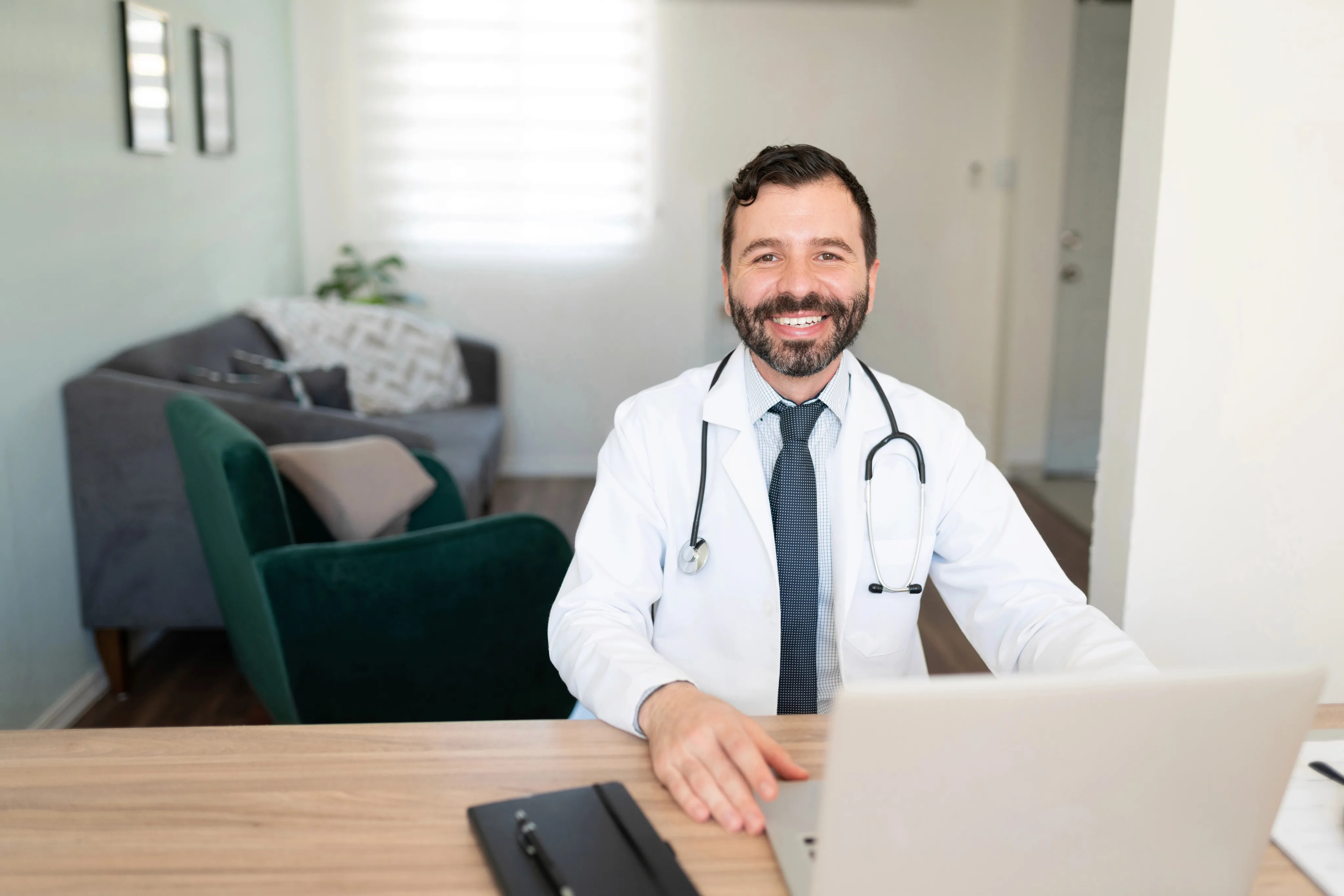A smiling male doctor wearing a white coat and stethoscope sits at a wooden desk with a laptop in a modern medical office with comfortable seating in the background.