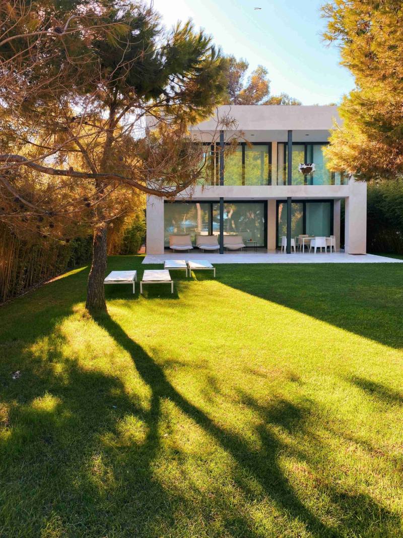 Modern white two-story house with glass walls, green lawn, lounge chairs, and pine trees in golden sunlight.