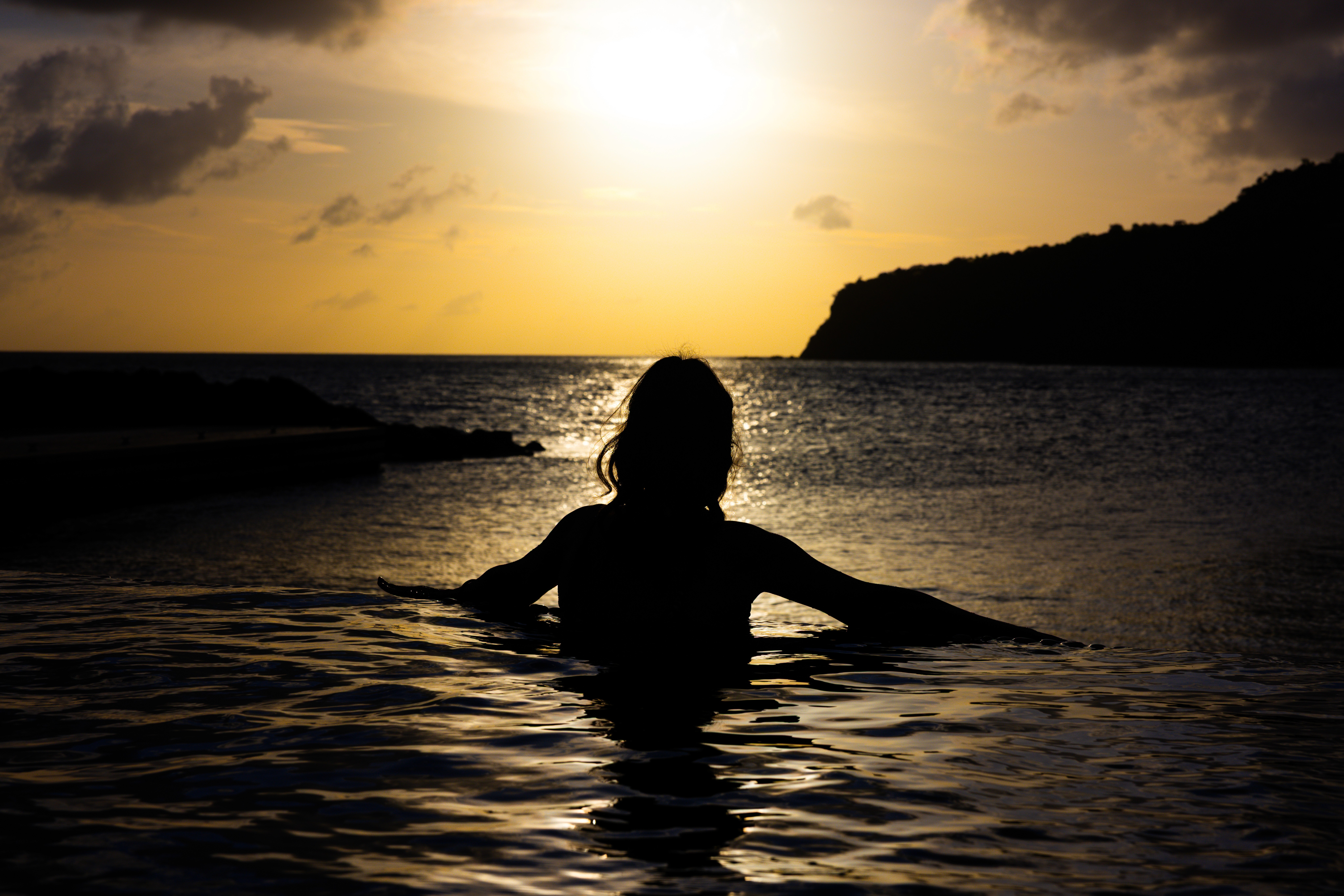 Person standing in calm ocean water at sunset, silhouetted against golden light