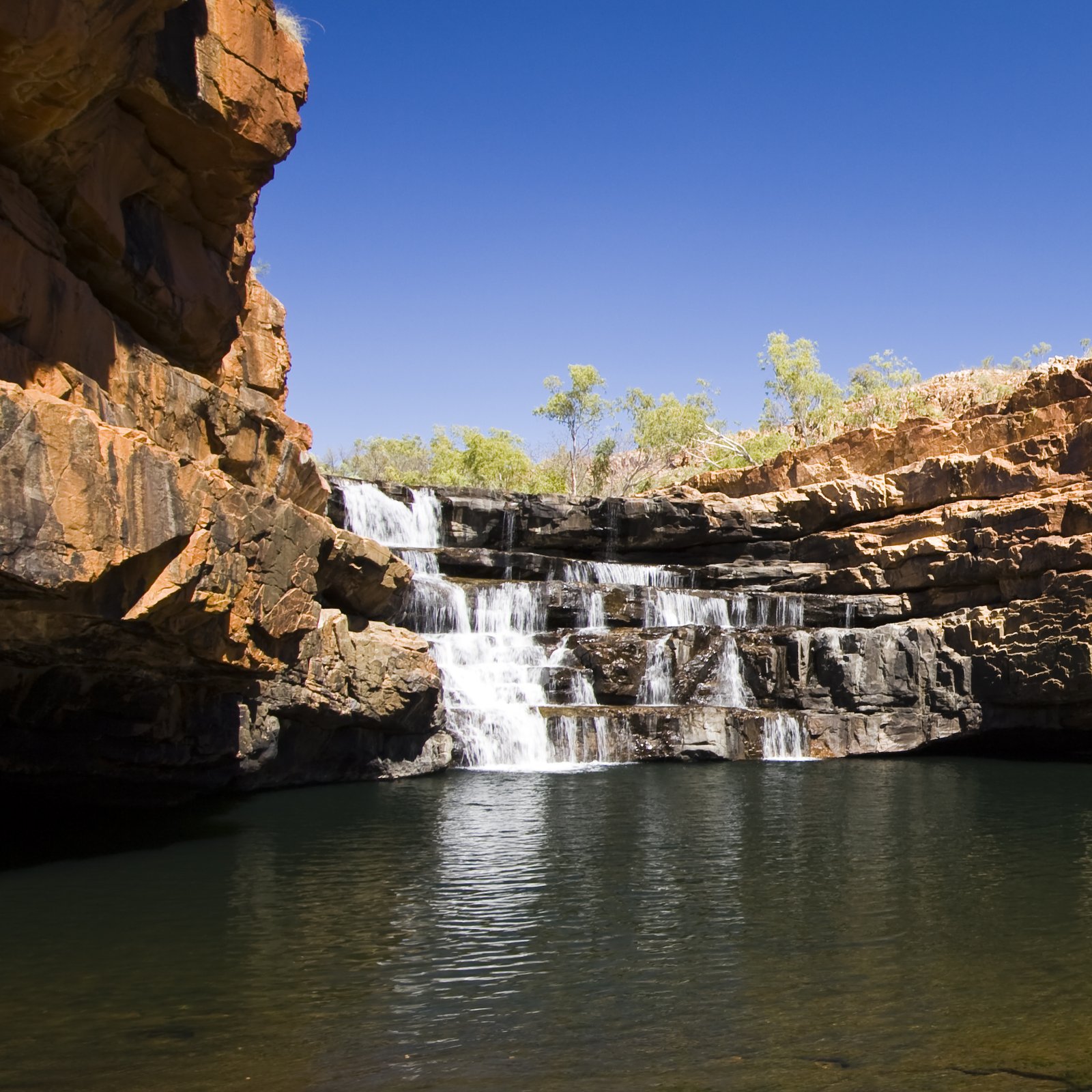 A layered red-rock gorge waterfall in the Kimberley, Western Australia, plunging into a clear plunge pool beneath sandstone cliffs.