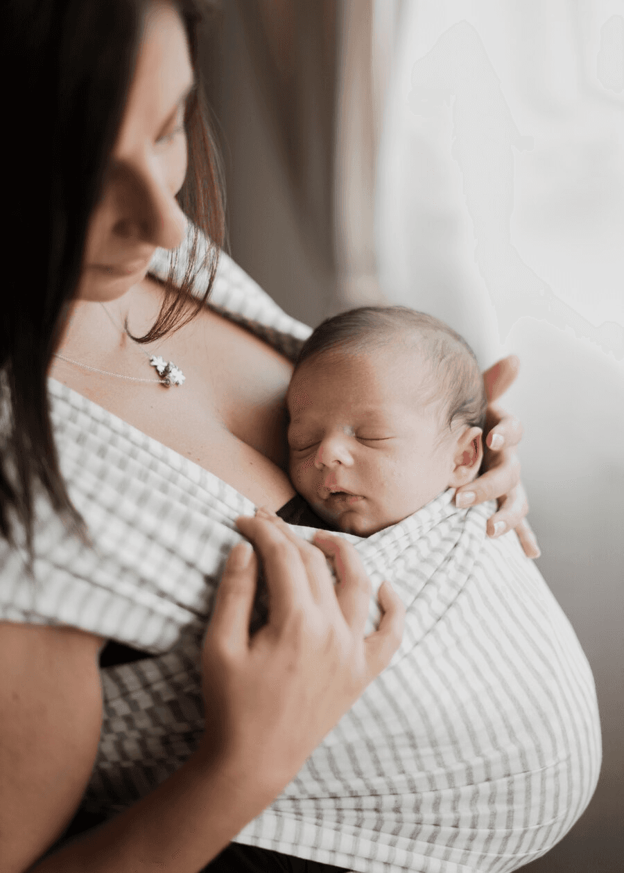 A woman gently cradles a sleeping newborn close to her chest, wrapped snugly in a striped fabric, illustrating tender postpartum care and intimate bonding.