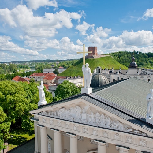 Rooftop view of Vilnius Cathedral with statues and a cross, lush greenery, and Gediminas Tower on a hill under a partly cloudy sky.