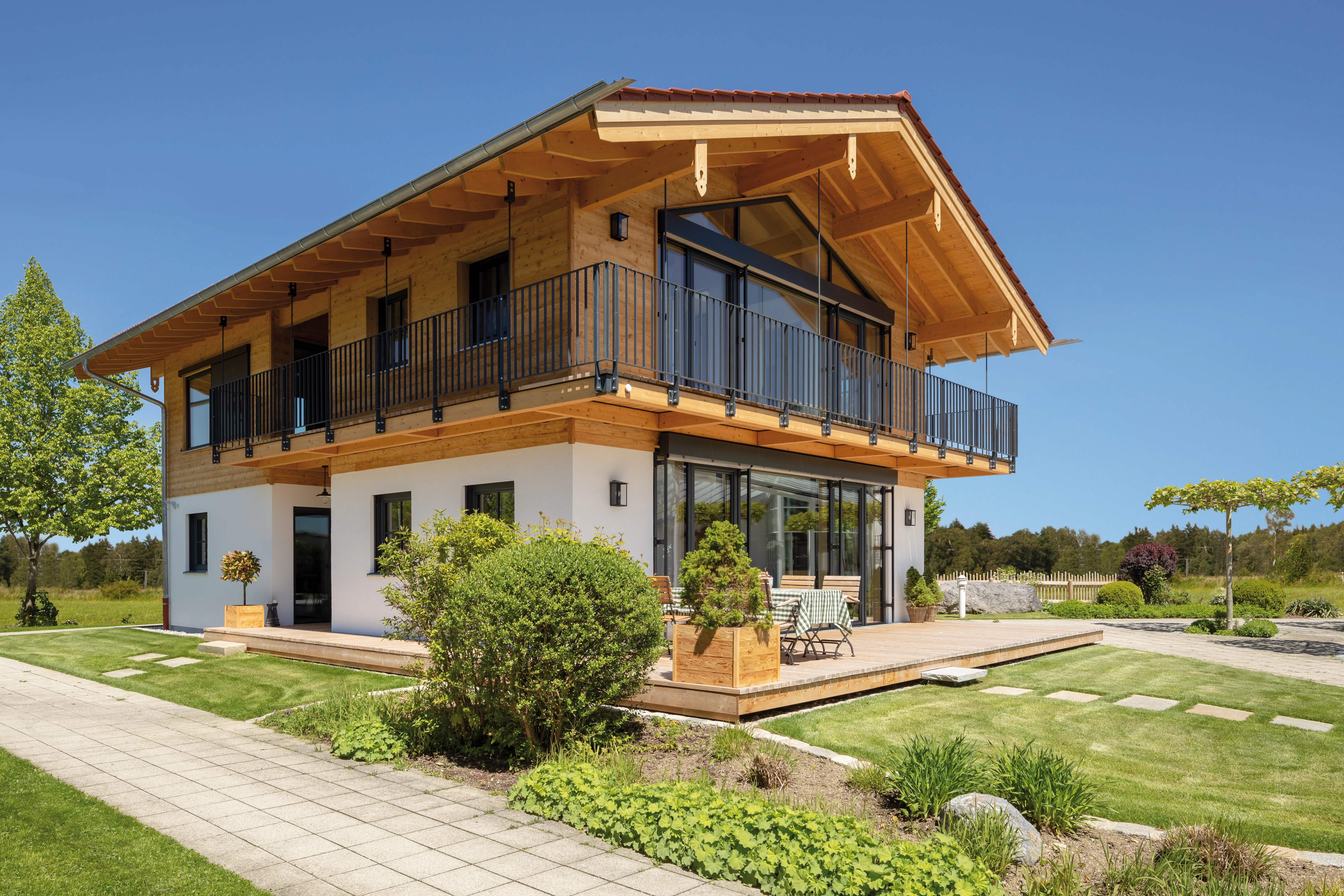 Modernes Holzhaus mit großzügigem Balkon, weißem Putz und weitläufiger Terrasse an einem sonnigen Tag.