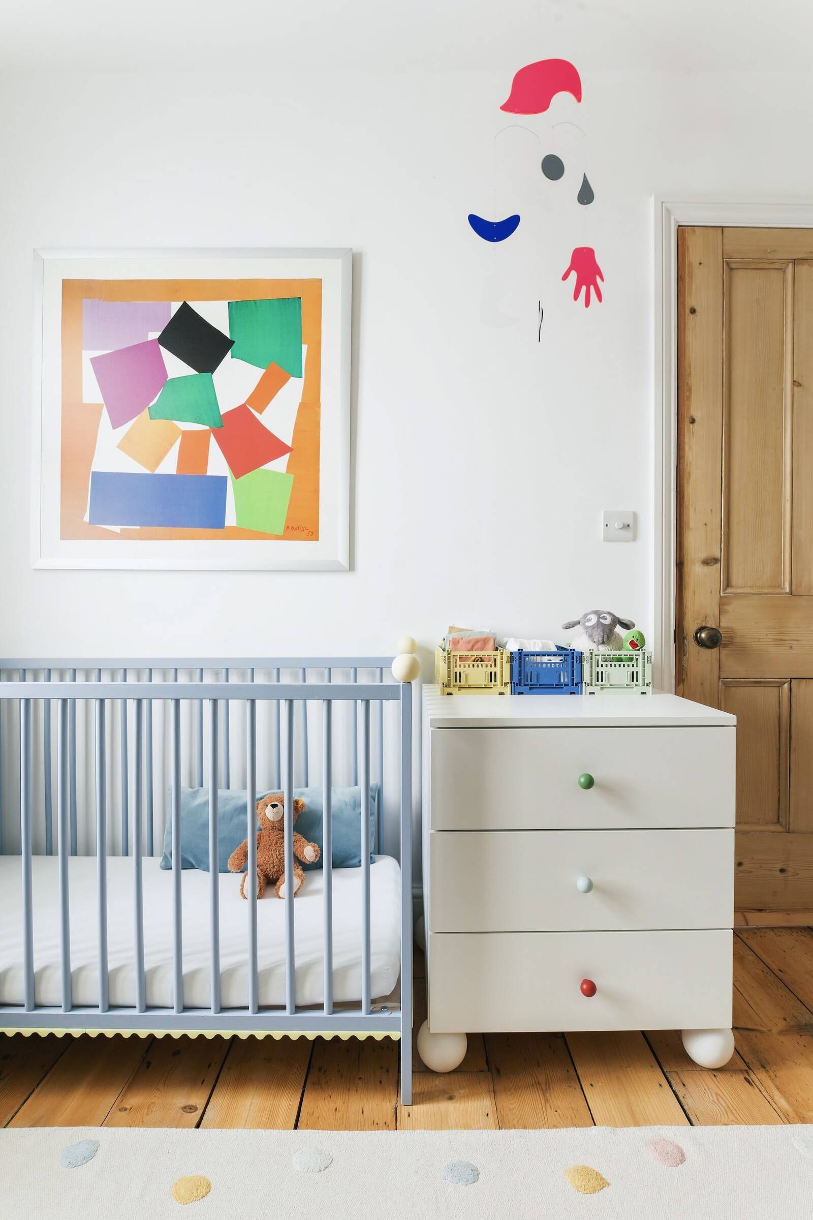 A baby's nursery with a light blue crib, a white dresser with colorful crate storage and stuffed toys, abstract artwork on the wall, and decorative wall hanging, all on a wooden floor with a polka dot rug.