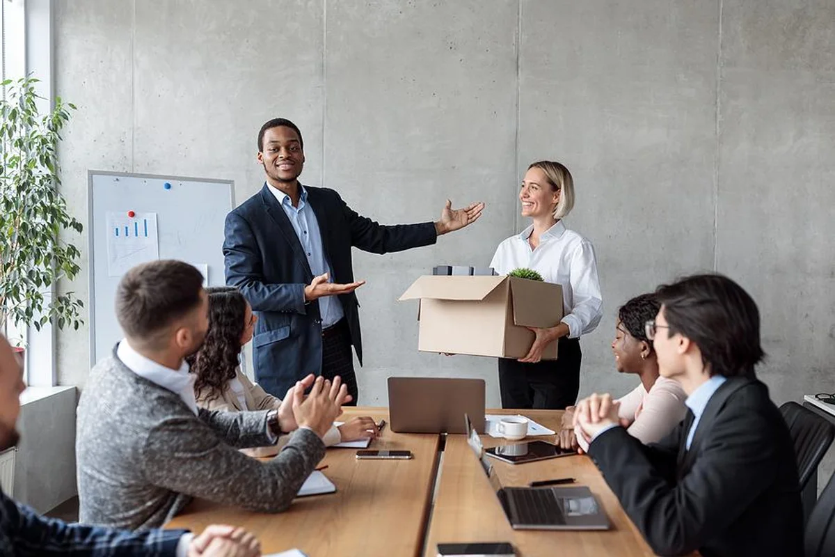 Man standing in a meeting room, saying goodbye to colleagues as he leaves for a new job. The atmosphere is emotional, with a handshake and smiles, while others sit or stand around the conference table.