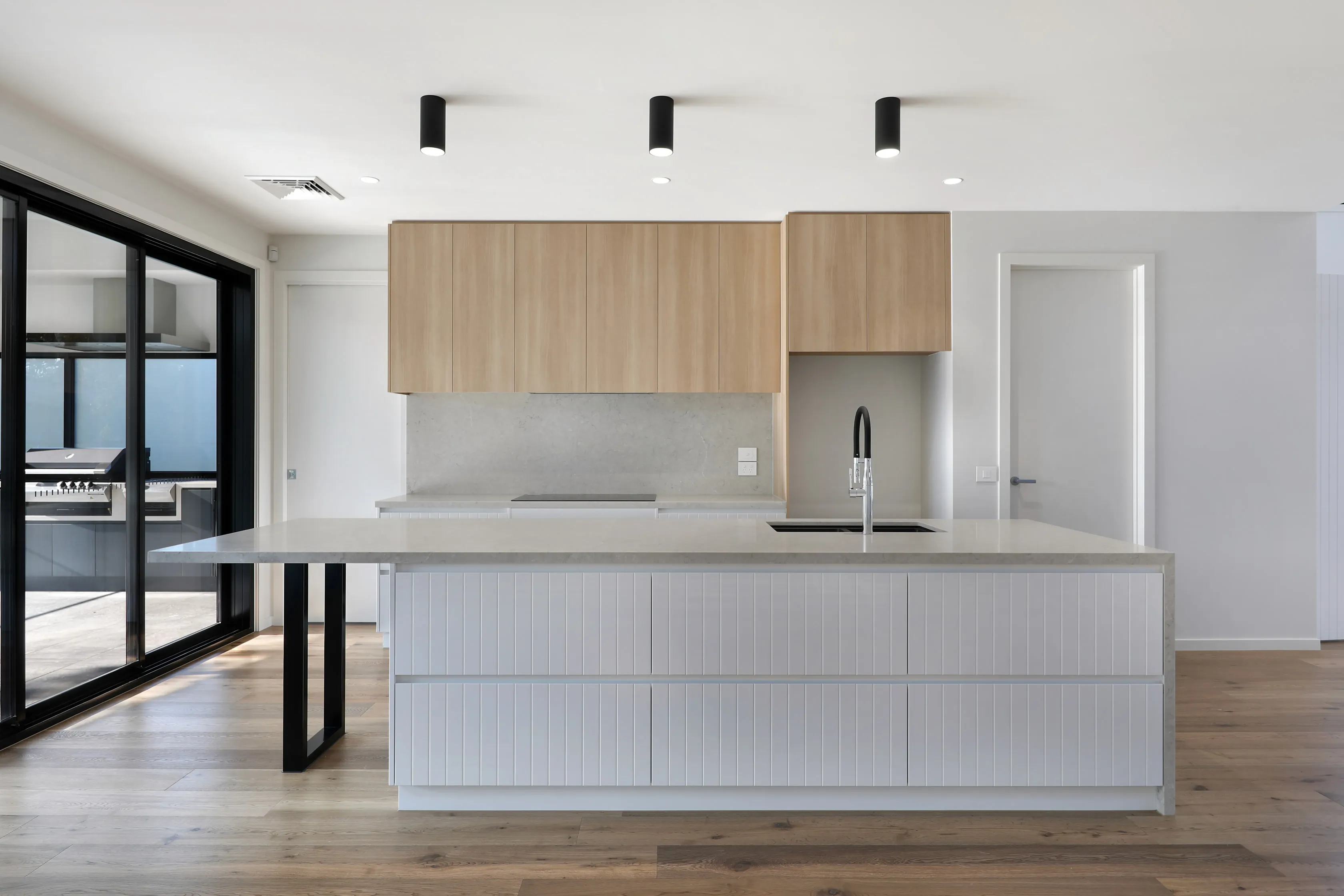 Kitchen with pale-oak overhead cabinetry, a fluted white island with grey stone benchtop on a black steel leg, black gooseneck tap, stone splashback and timber floors, beside black sliding doors to the alfresco