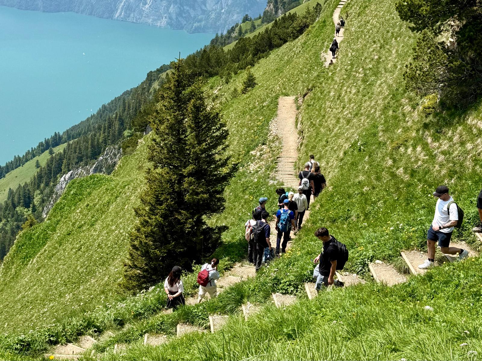 A group of hikers following a narrow mountain trail above a turquoise alpine lake.