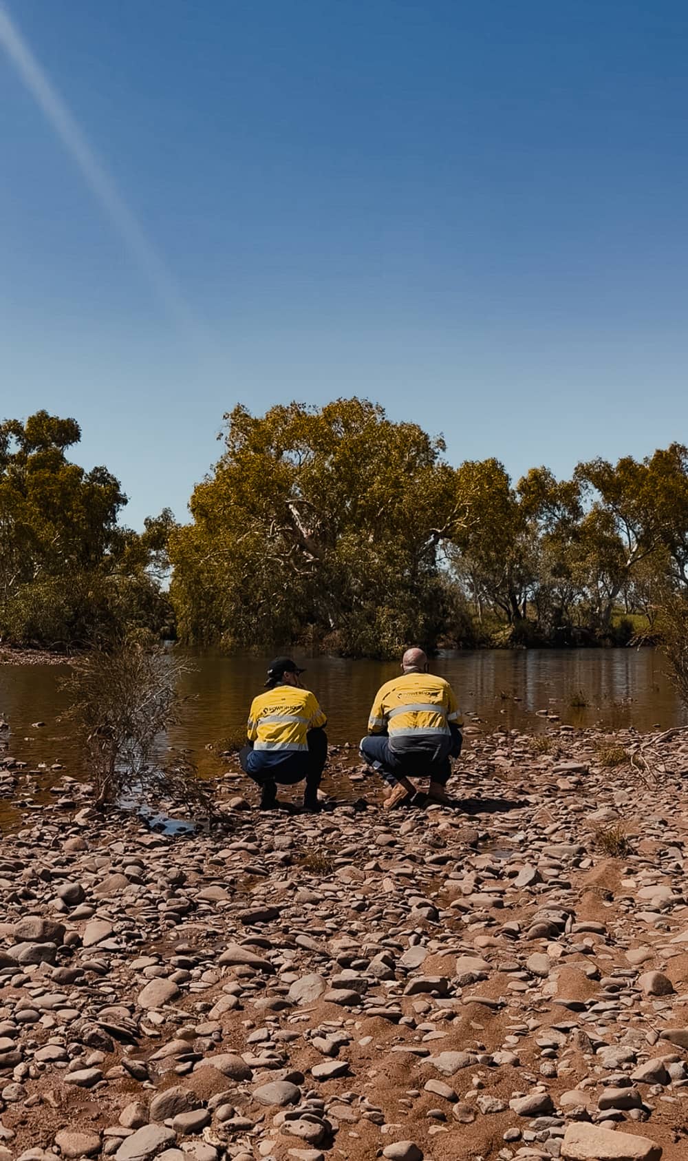 Two Powerhouse Renewables Group technicians in yellow high-vis workwear crouch by a rocky riverbank in remote Australia.