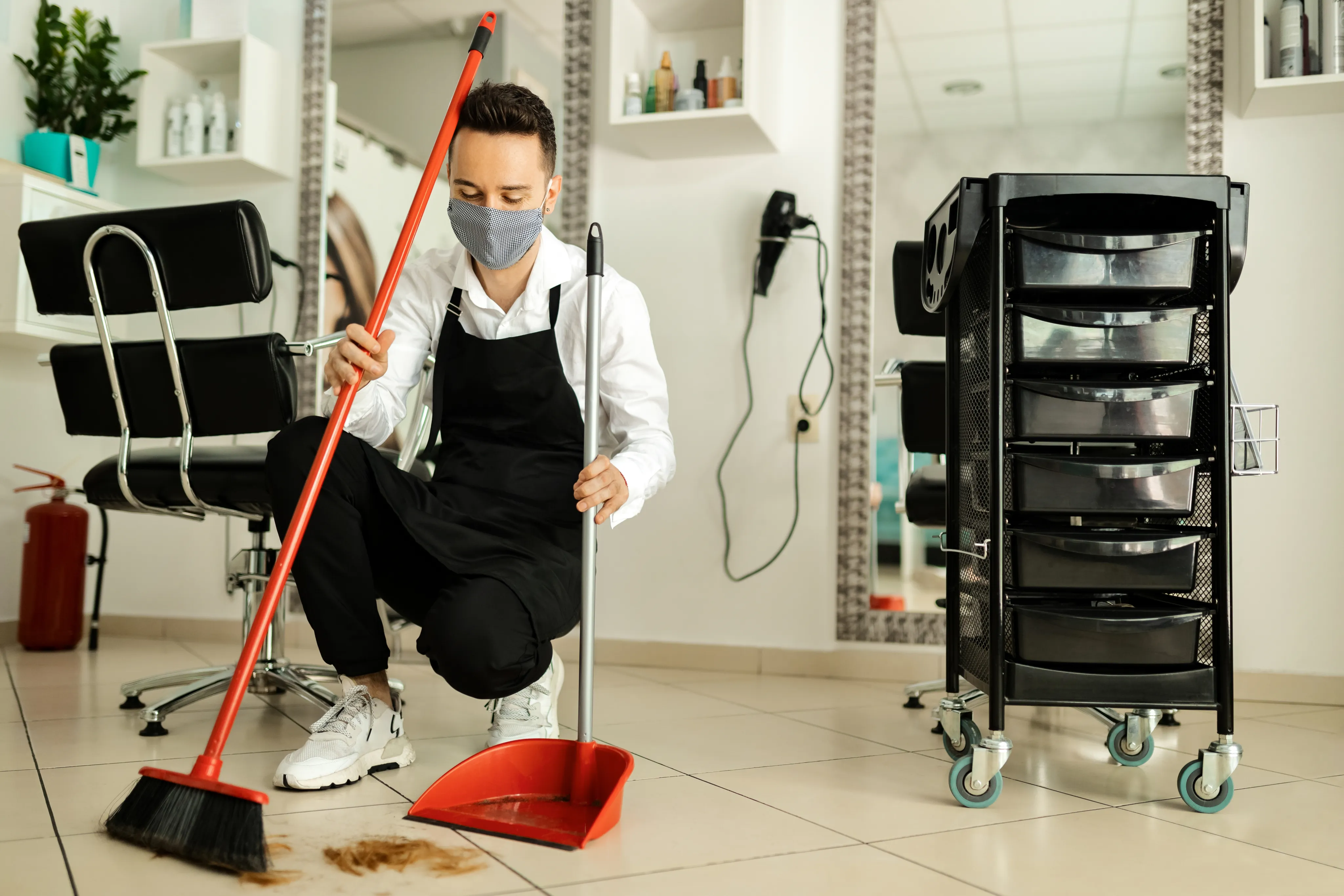 A male cleaner in a face mask and apron sweeping the floor of a professional hair salon using a red dustpan and broom.
