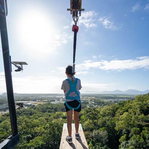 一个人穿着安全带站在平台上，准备搭乘滑索飞越一片森林，背景中可以看到小镇和山脉。