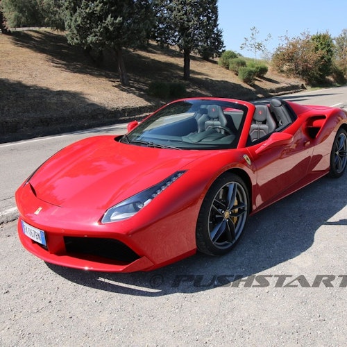 A red convertible sports car parked on the side of a road near a grassy hill with trees.