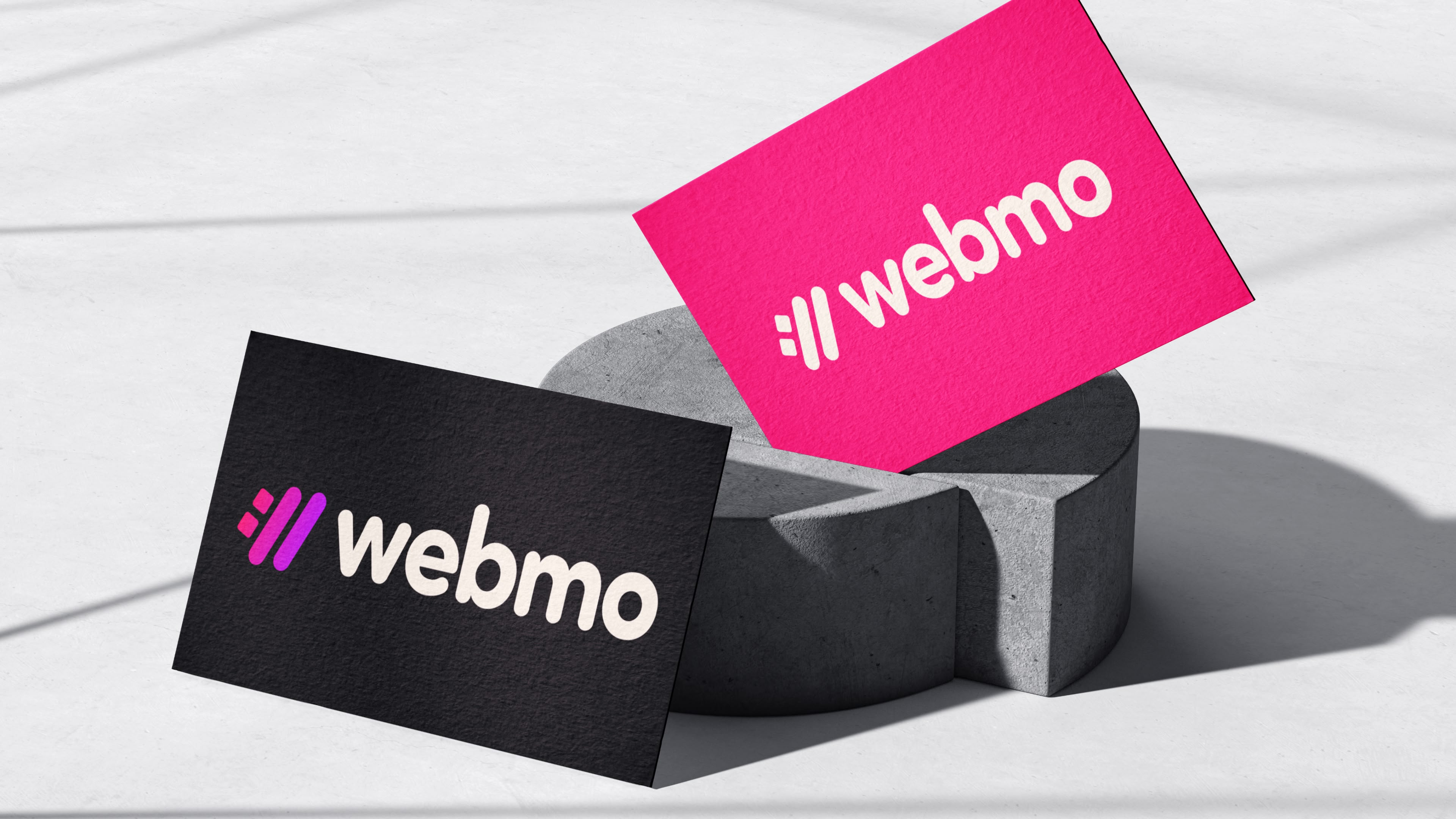 A business card design mockup with one front having black and the Webmo logo icon in a gradient of pink to purple and the Webmo logo type in a white color. Another business card mockup front with magenta color and the Webmo logo icon. logo type in white. One of the magenta cards is wedged in between a very interesting cement sculpture, while the black business card front is rested against the cement sculpture. They are sitting on a modern white marble tabletop with contrast lighting. 