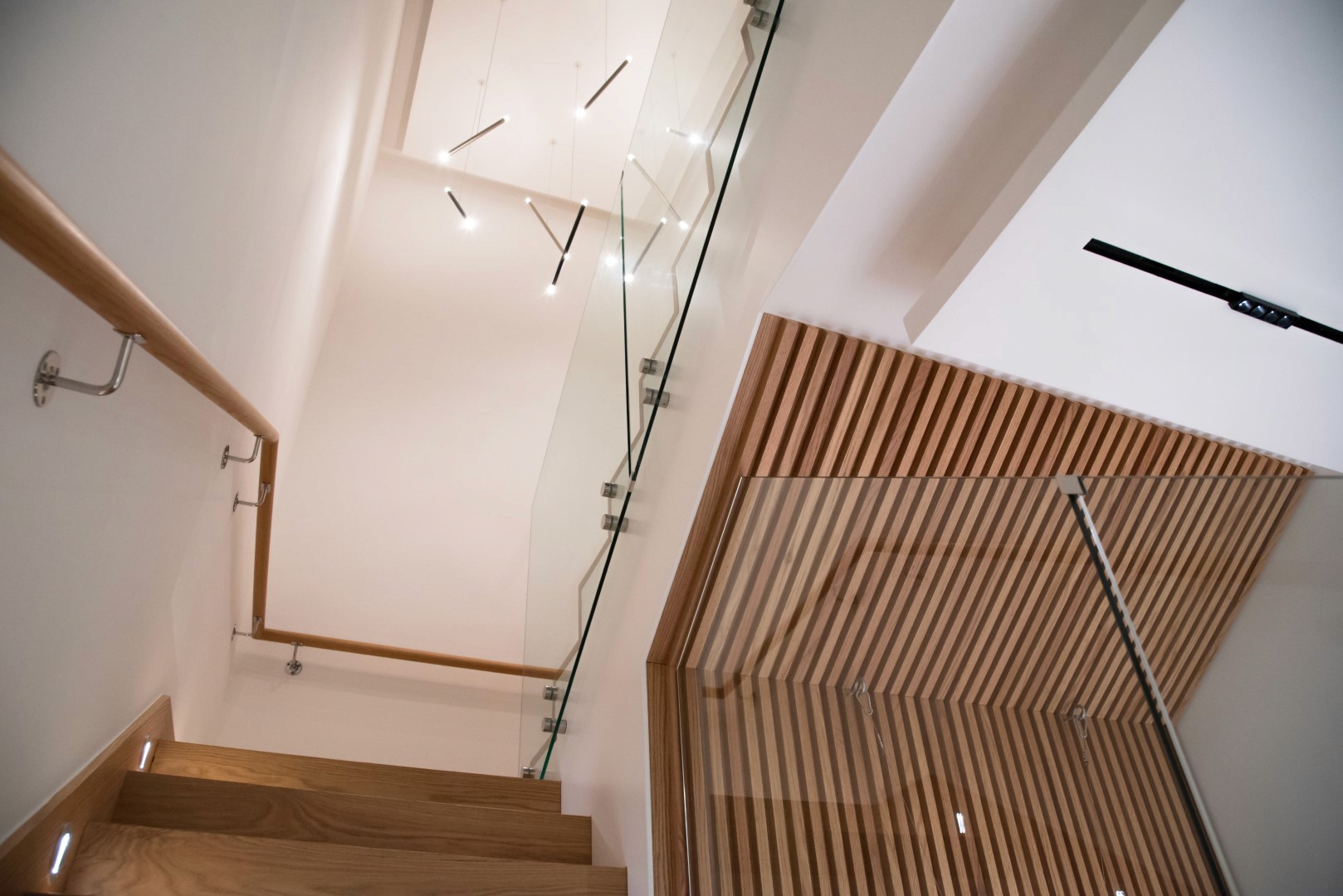Modern staircase featuring wooden steps and railing, glass balustrade, and sleek overhead lighting in a minimalist interior design setting.