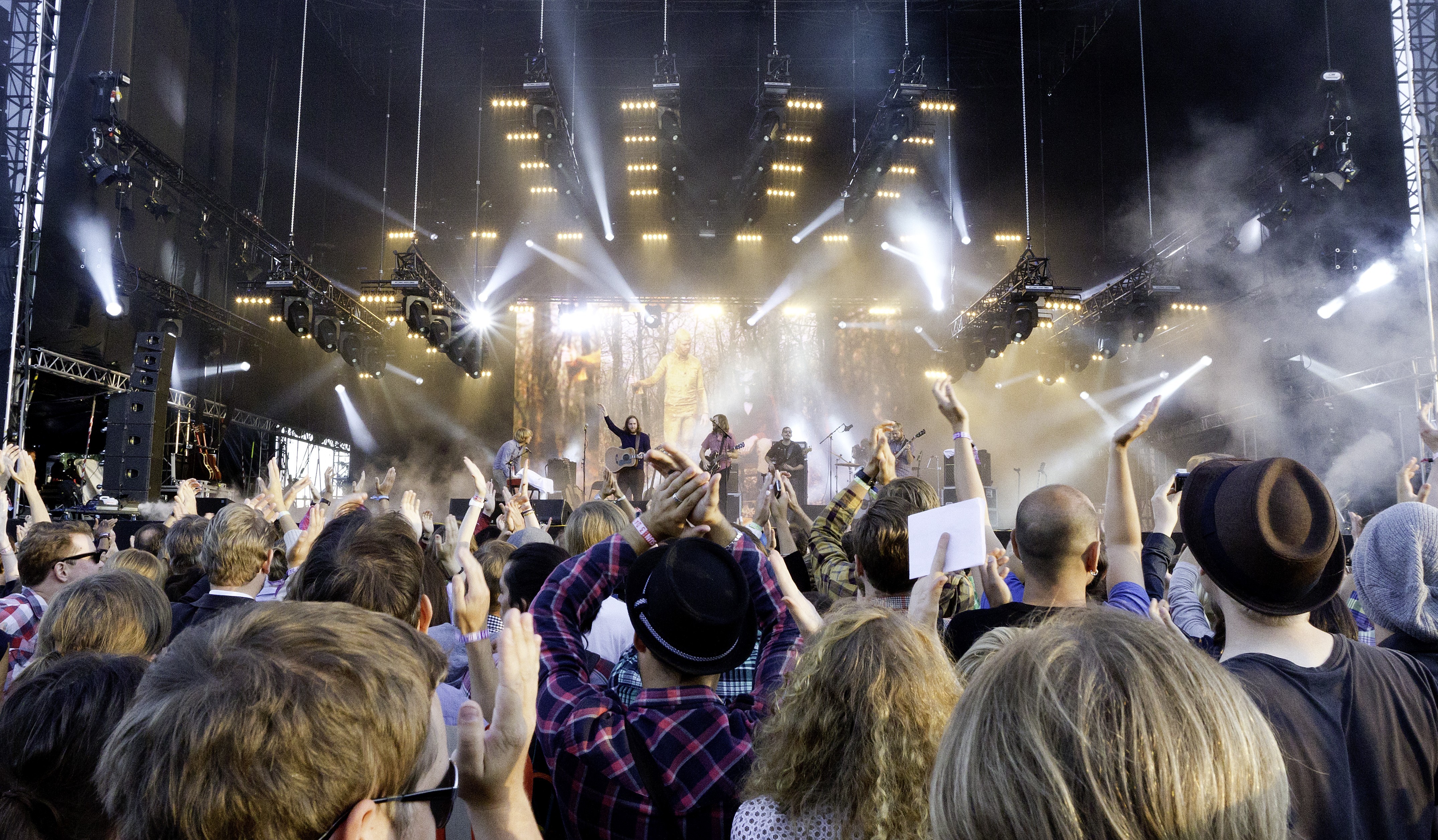 Crowd cheering at an outdoor music festival as a band performs on a brightly lit stage.