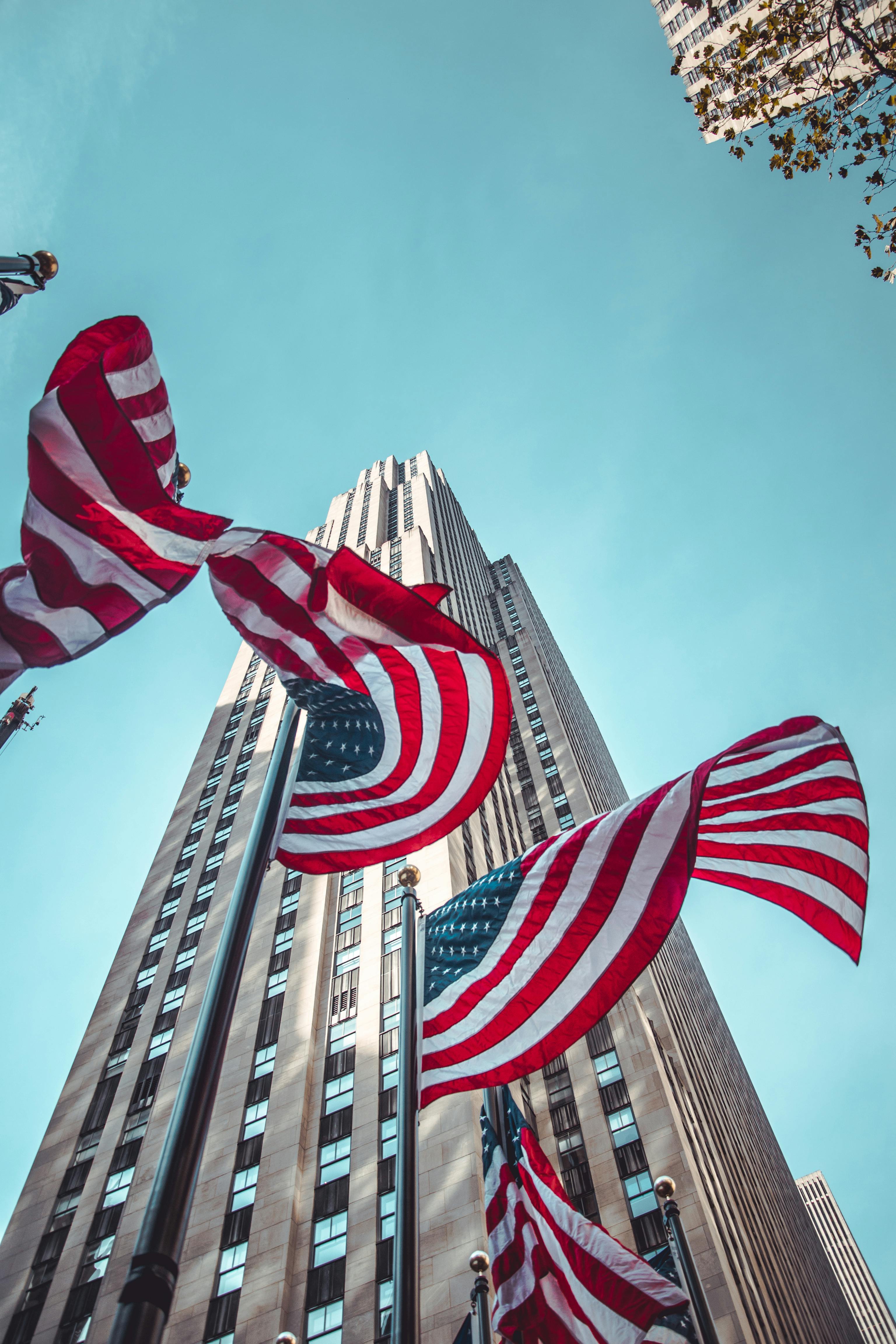 A low-angle shot of the Rockefeller Center in New York City. Several American flags flutter in the foreground, framing the tall Art Deco skyscraper against a clear blue sky.