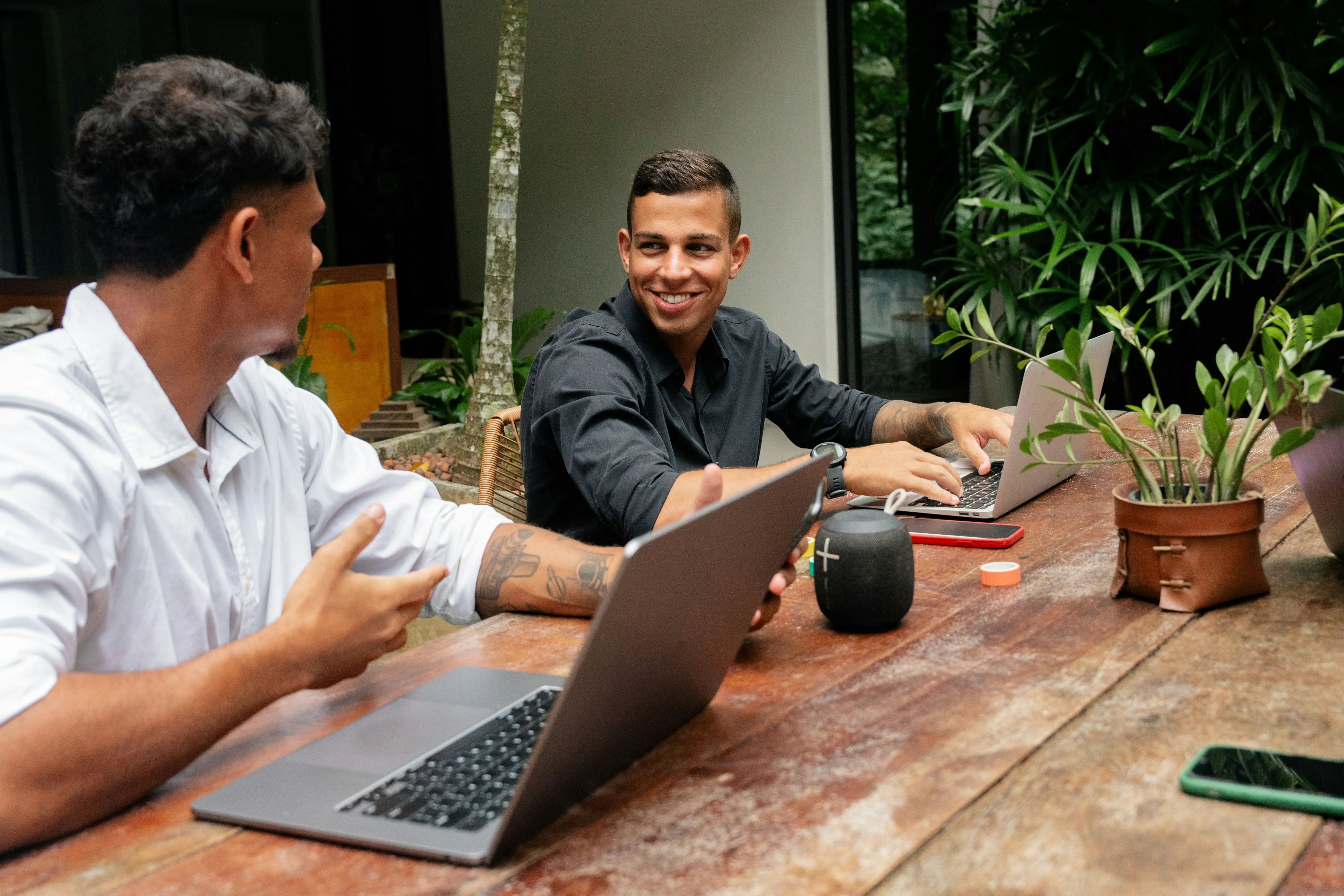 Two men sit at a table with laptops, discussing ideas in a modern workspace filled with plants.