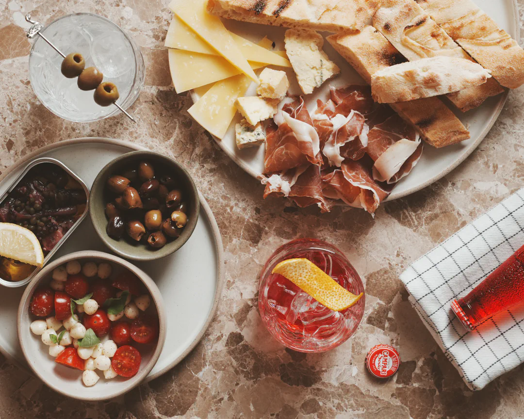 Sharing board of cheese, cured ham, flatbread, olives and tomatoes with a spritz on a marble table