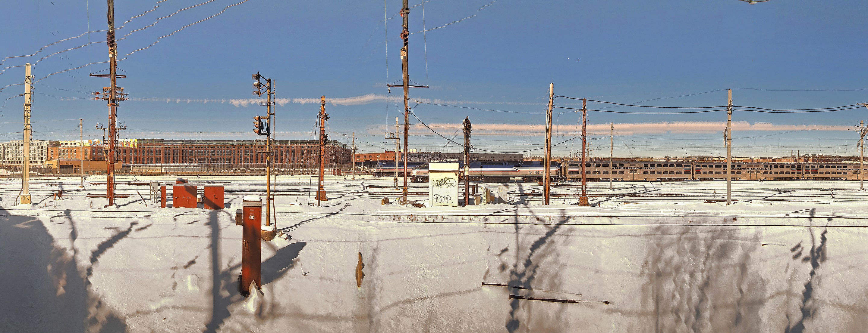 Panoramic photograph of a snow-covered train yard with power poles and industrial buildings under a clear blue sky. Subtle distortions stretch the lines of cables and shadows, transforming the static scene into a meditation on stillness and movement.