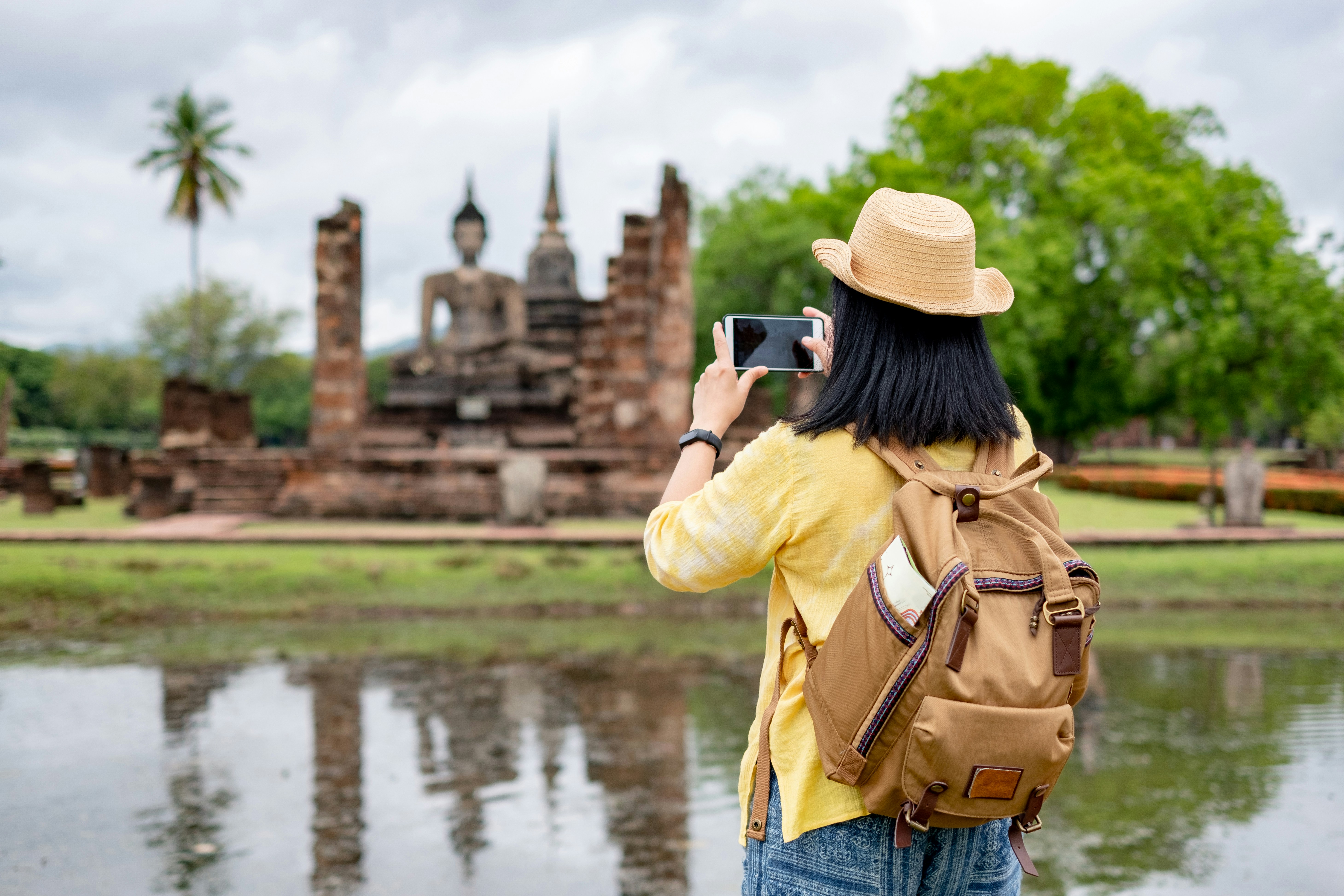 A traveler wearing a straw hat and backpack takes a photo with a smartphone of an ancient stone Buddha statue and temple ruins across a reflective pool in a lush, green historical park.
