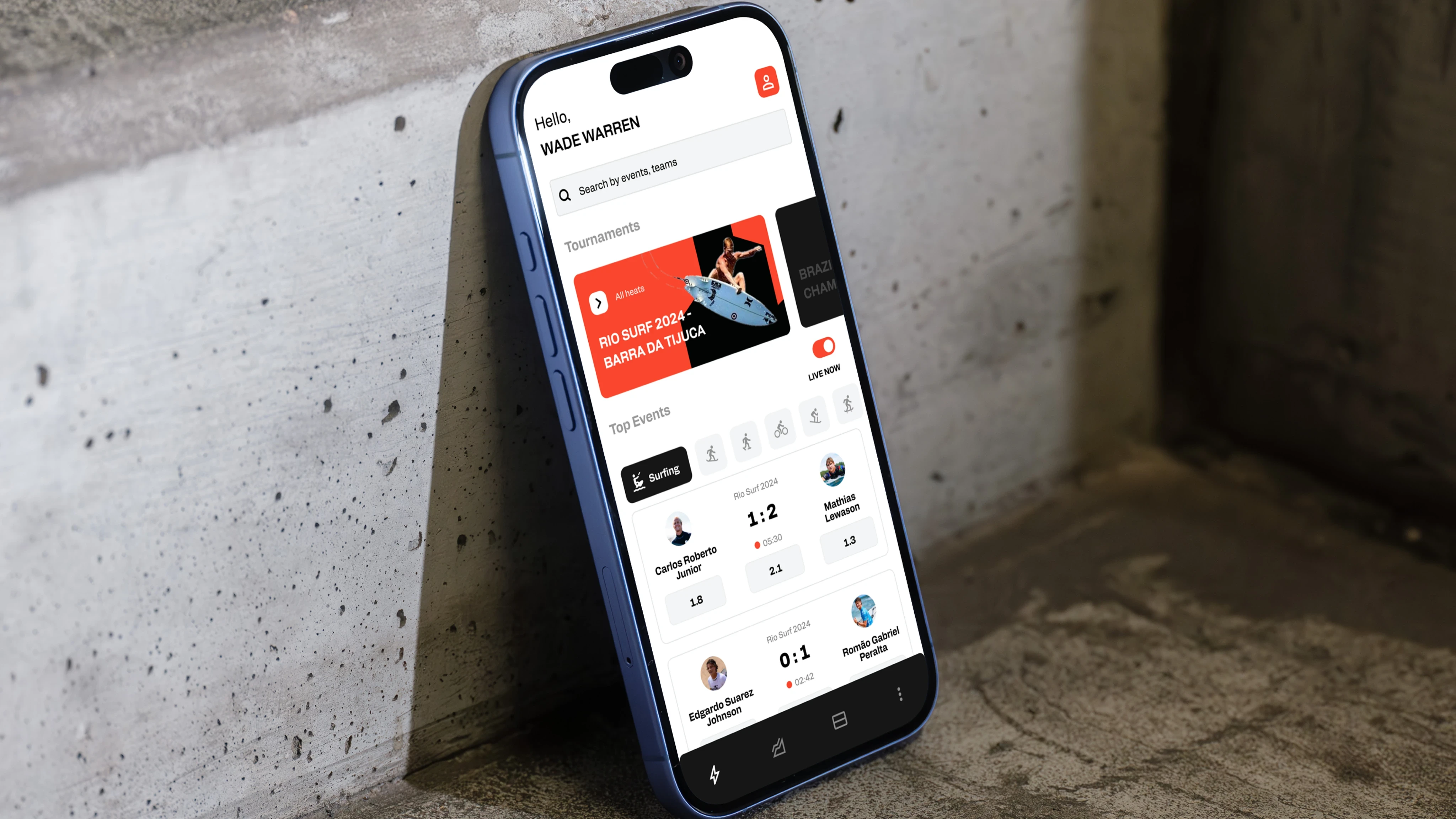 Smartphone with a sports event app open, displaying surfing tournament scores and match details, leaning against a concrete wall.