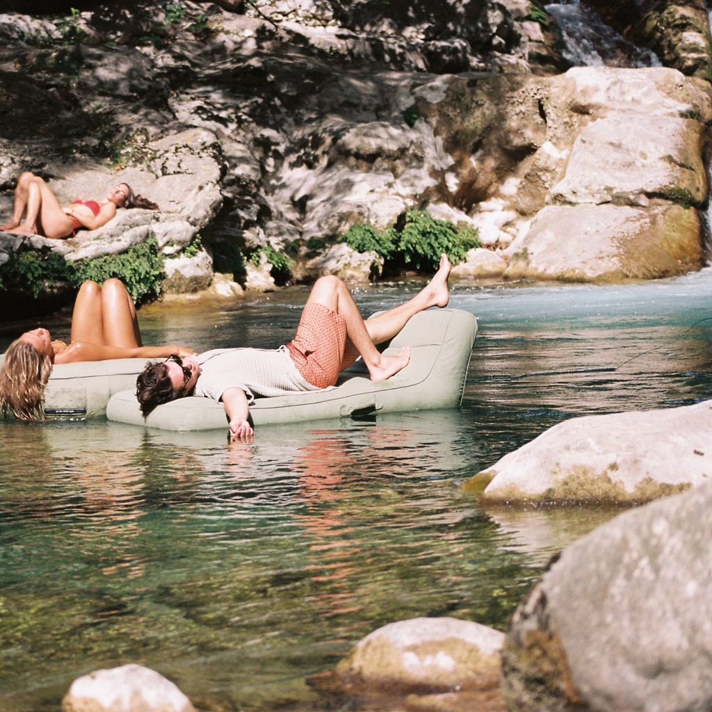 A group of people relax in the river: two float on Oliver James Lilos Single in Leaf, sunlight on LiloCover upholstery, one sunbathes nearby.