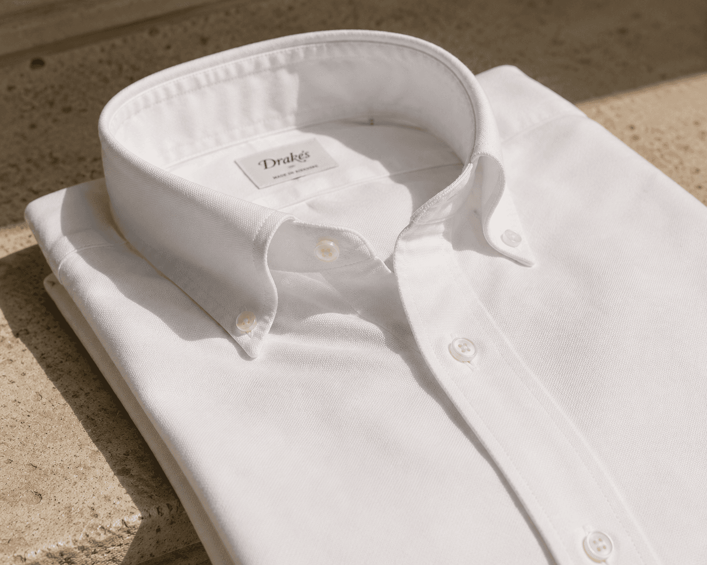 A close-up shot of a neatly folded crisp white men's oxford cloth button-down shirt (OCBD) resting on a textured stone surface. The focus is on the collar construction, showing a beautiful, soft collar roll with white buttons securing the points. The inside collar label reads "Drake's", with a smaller tag below it that says "MADE IN SOMERSET".
